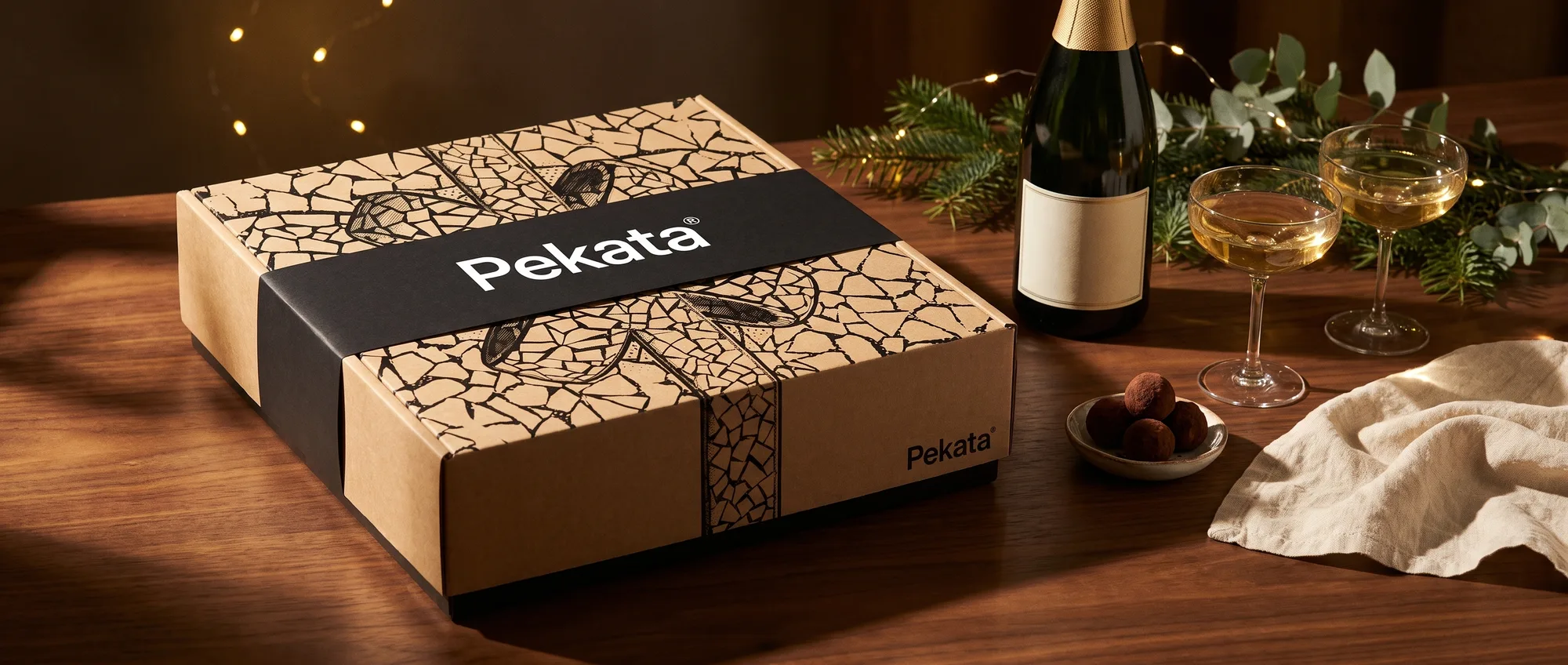 Caixa de Natal Pekata fechada com faixa preta ao lado de uma garrafa de espumante e dois copos sobre uma mesa de nogueira